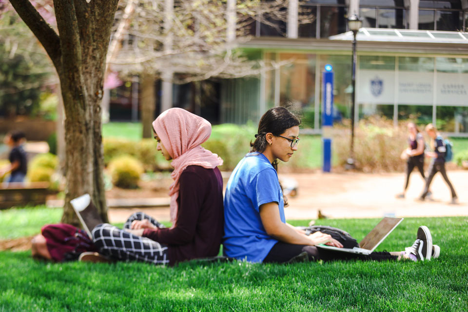 Two slu students sit back to back on the ground under a tree on the SLU Quad.