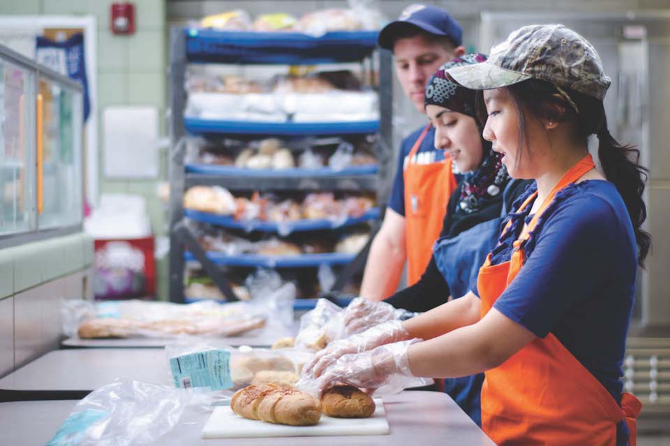 Three students are shown in profile, wearing aprons, working with bread on a kitchen counter.
